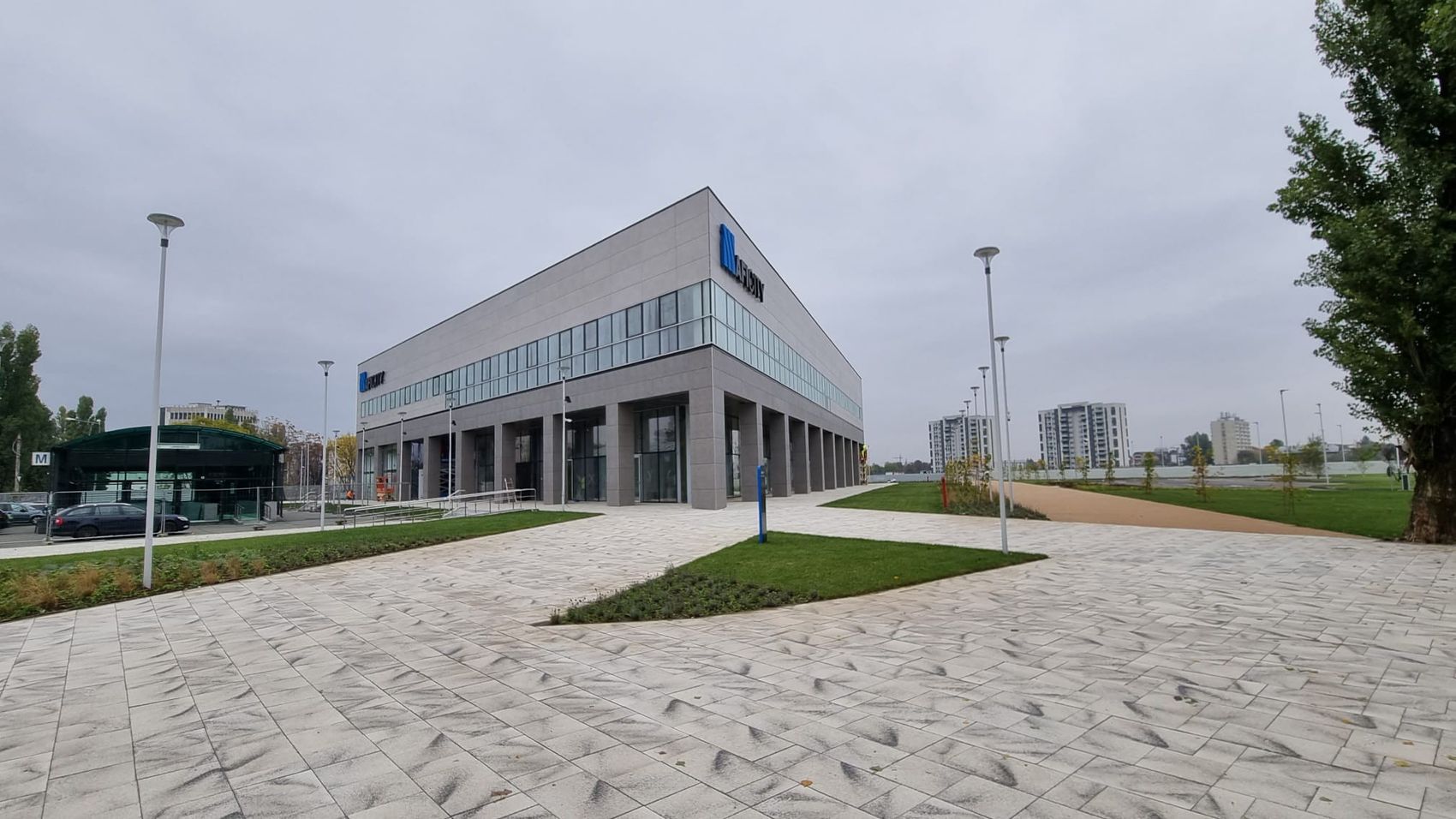 Clădirea comercială AFI City din Bucureștii Noi, cu fațadă gri și o bandă de vitraje, înconjurată de alei pavate și gazon nou, sub cer înnorat