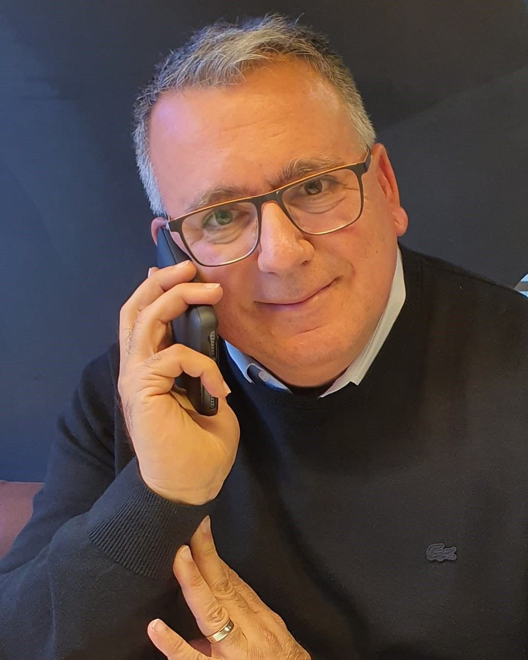 Portretul lui Yossi Moran, cu ochelari și pulover închis la culoare, vorbind la telefon pe un fundal închis